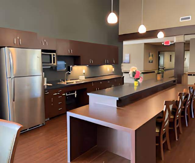 Communal kitchen and dining area with a long island counter, bar stools, stainless steel refrigerator, and pendant lights.