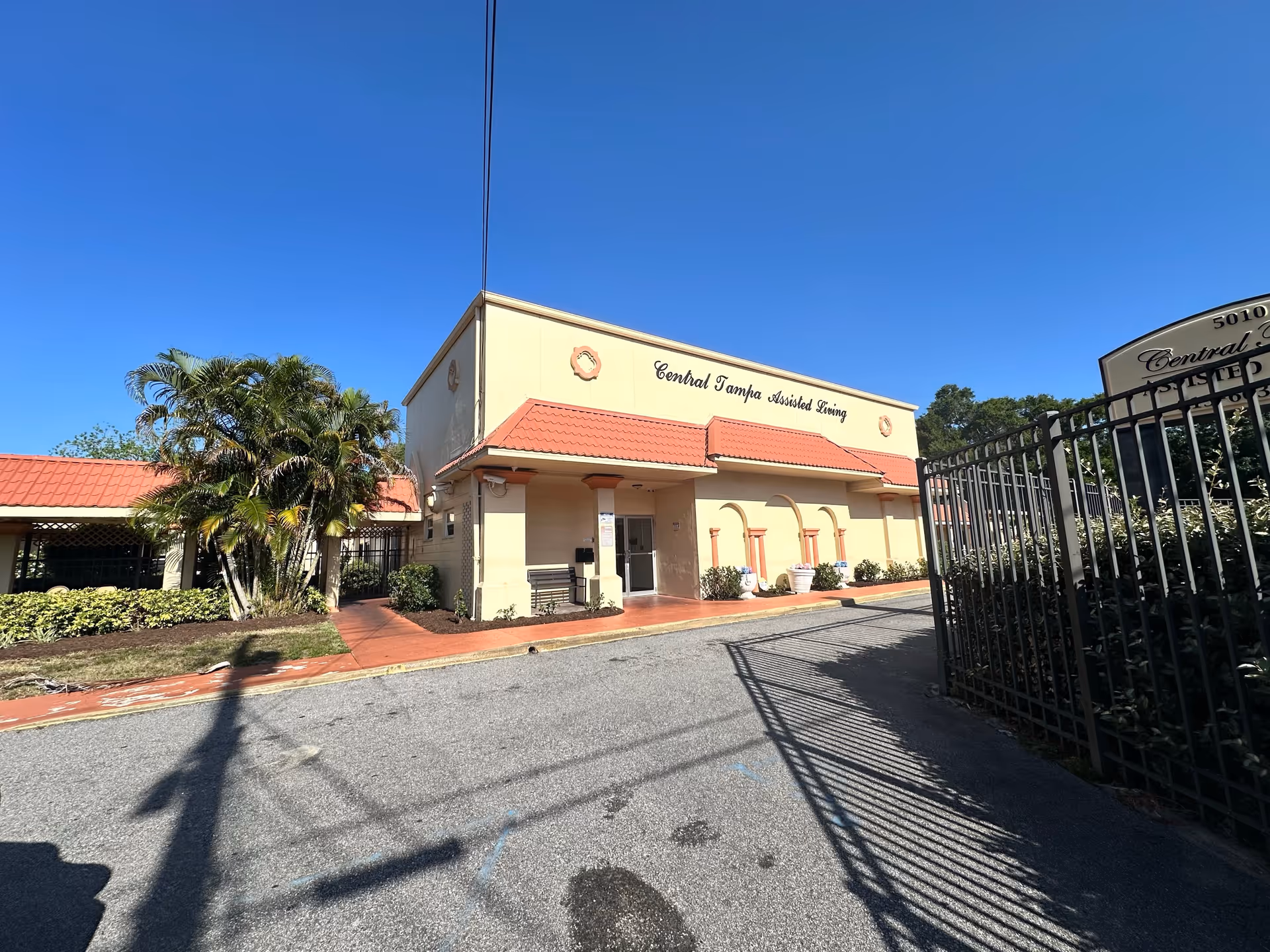 Exterior view of a beige building with a red tiled roof labeled 'Central Tampa Assisted Living' under a clear blue sky. There are palm trees and shrubs near the entrance, a paved driveway, and a black metal fence on the right side.