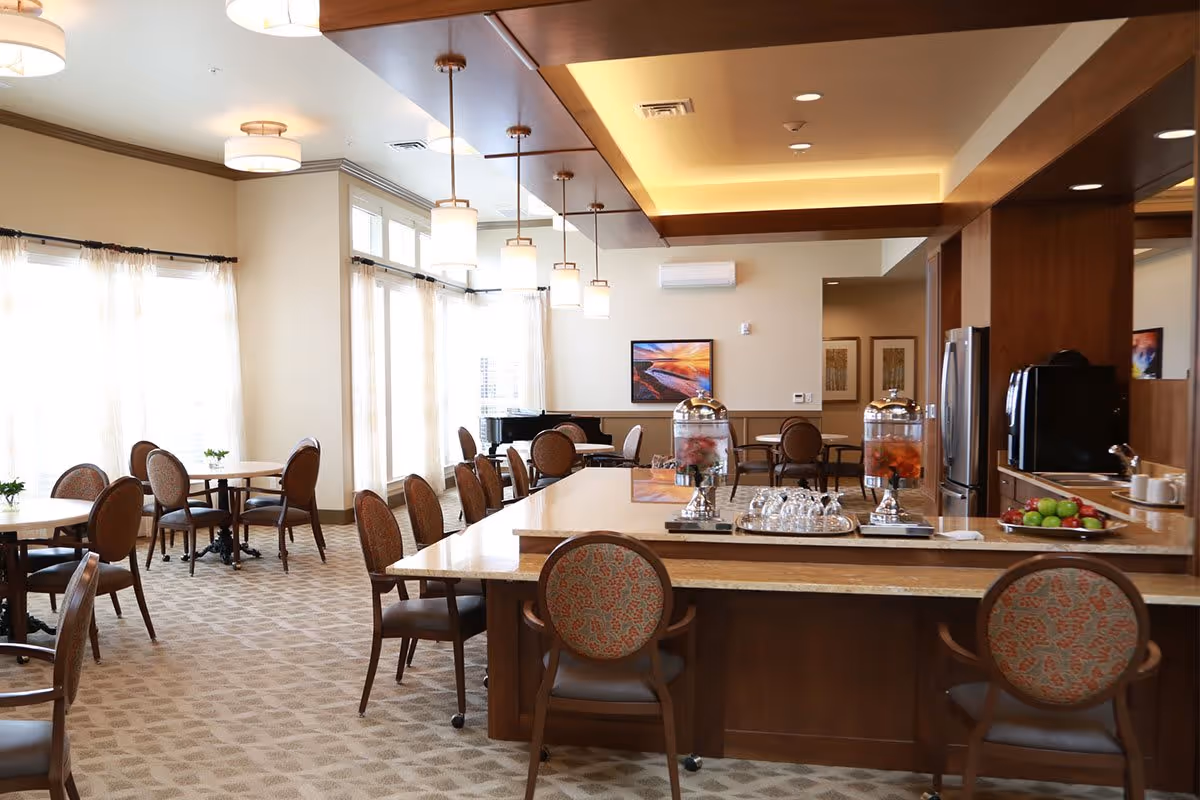Spacious, well-lit communal dining room with round tables and a central island offering beverages and fruit.