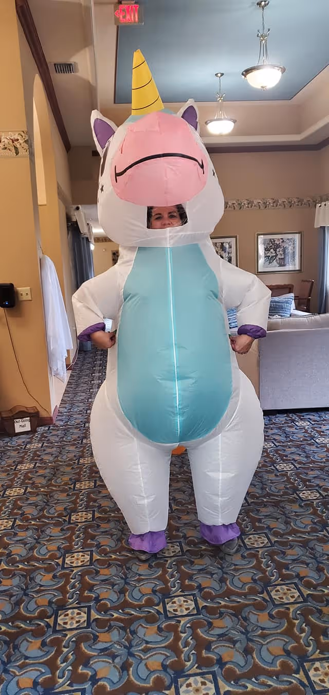 Person standing indoors wearing a large inflatable unicorn costume with a yellow horn, pink snout, and teal belly. The room has patterned carpet, beige walls, and ceiling lights. There is a couch and framed pictures in the background.