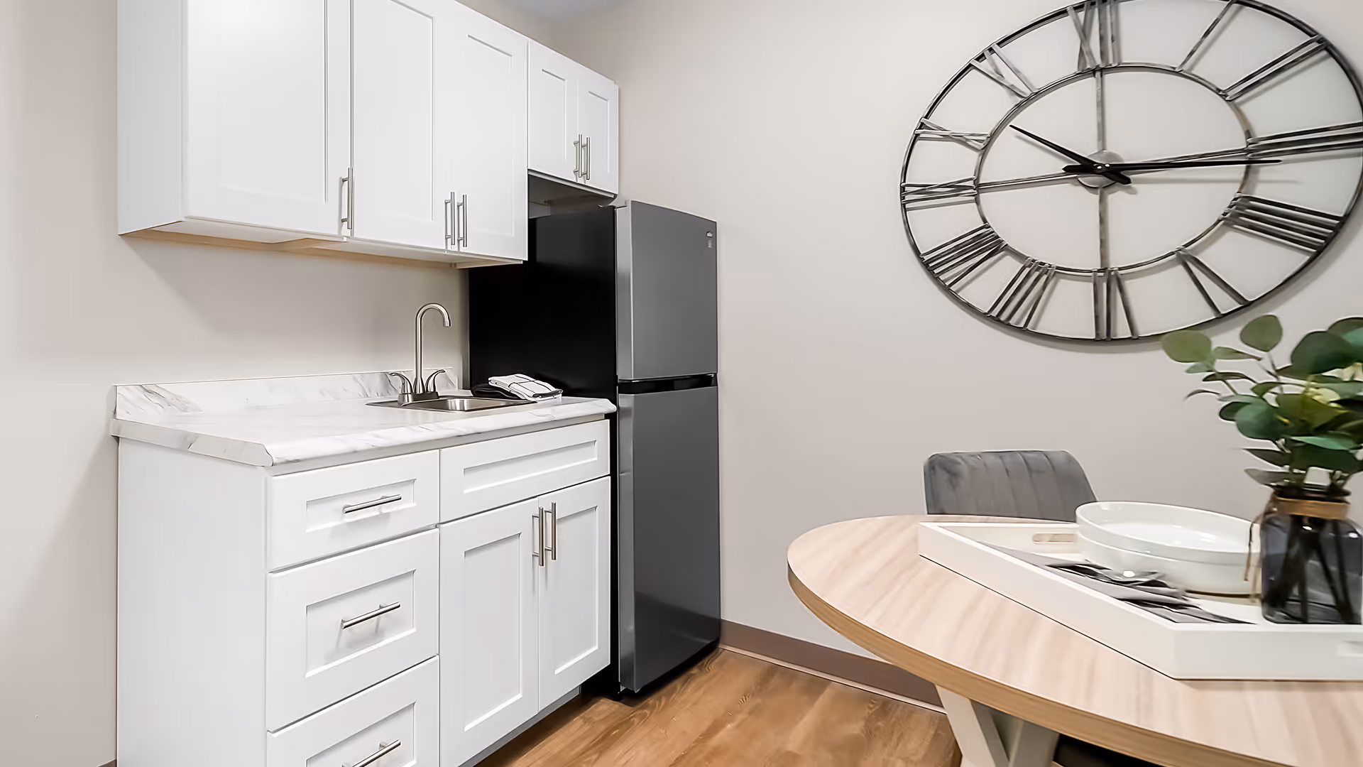 Small kitchenette with white cabinets, marble-look countertop, stainless mini-fridge, round dining table and an oversized wall clock on the wall.