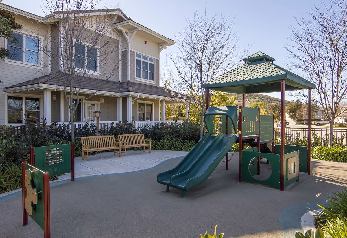 Outdoor playground area with a green double slide and climbing structure, surrounded by trees and bushes. Two wooden benches are placed on a paved area near the playground. In the background, there is a two-story residential-style building with multiple windows and a porch.