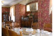A dining room with a wooden dining table set with cups, glasses, and plates. The room features floral wallpaper in red tones, wooden chairs, a wooden cabinet with decorative items, and windows with sheer curtains allowing natural light in.