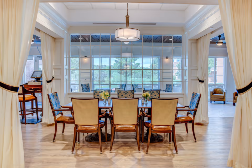 A bright and elegant dining room with a wooden table surrounded by eight upholstered chairs. The room features large windows with a view of greenery outside, light-colored curtains tied back, and a modern ceiling light fixture hanging above the table. The floor is wooden, and there are additional seating areas visible through the windows and open spaces.