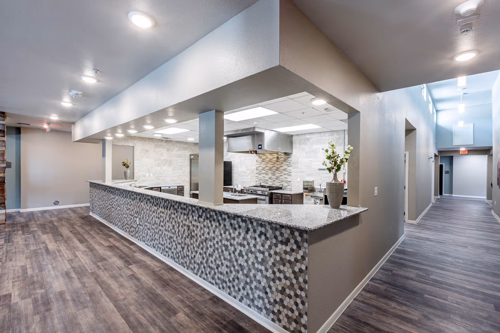 Interior view of a modern kitchen area in a senior living facility with a long granite countertop and hexagonal tile backsplash. The kitchen is open to a hallway with wood-style flooring and neutral-colored walls. There are ceiling lights and a vase with flowers on the countertop.