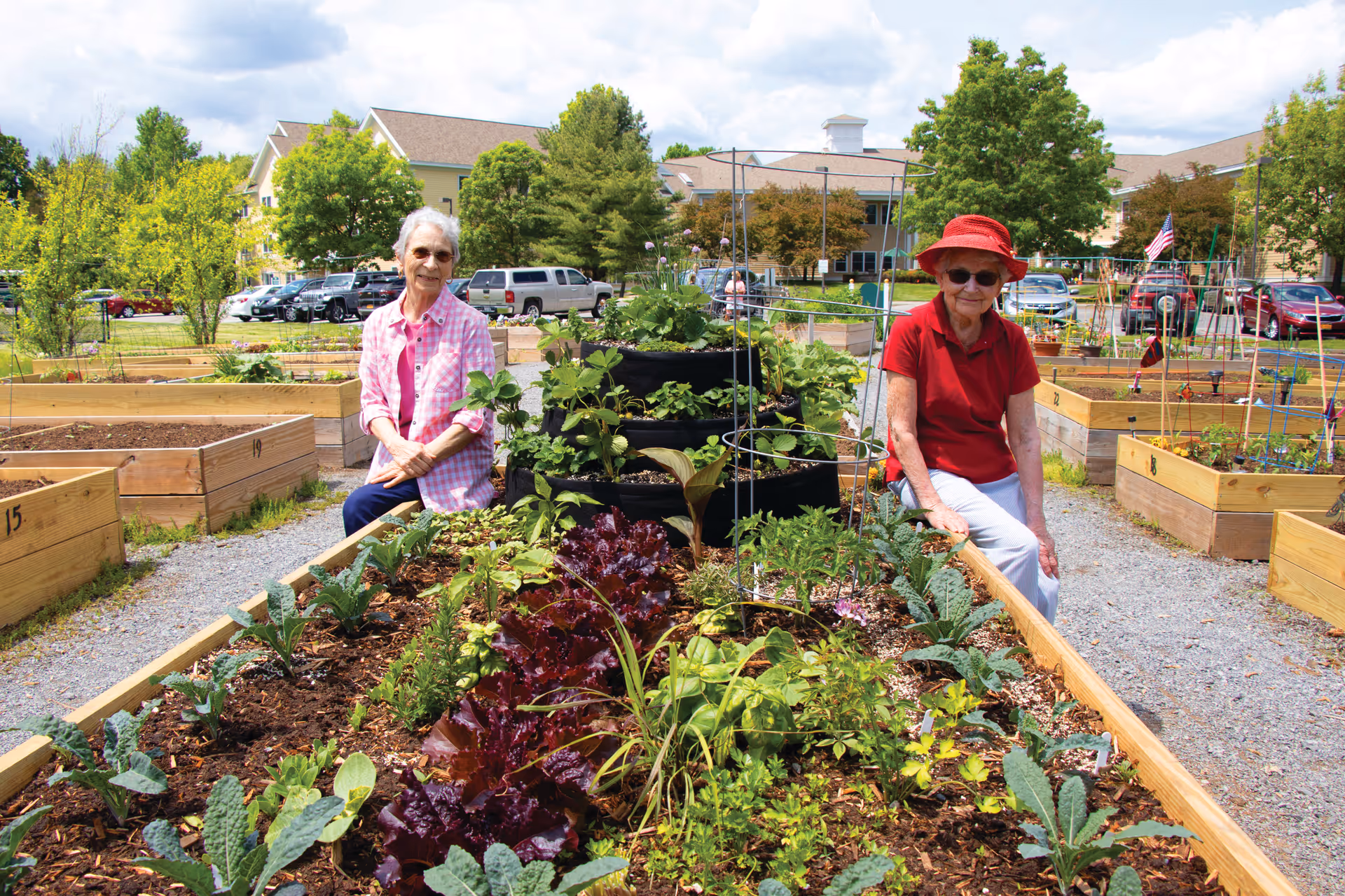 Two elderly women sitting on the edges of raised garden beds filled with various plants and vegetables in an outdoor community garden area. One woman is wearing a pink checkered shirt and glasses, and the other is wearing a red shirt and red hat. Behind them are more raised garden beds, parked cars, trees, and a large building under a partly cloudy sky.