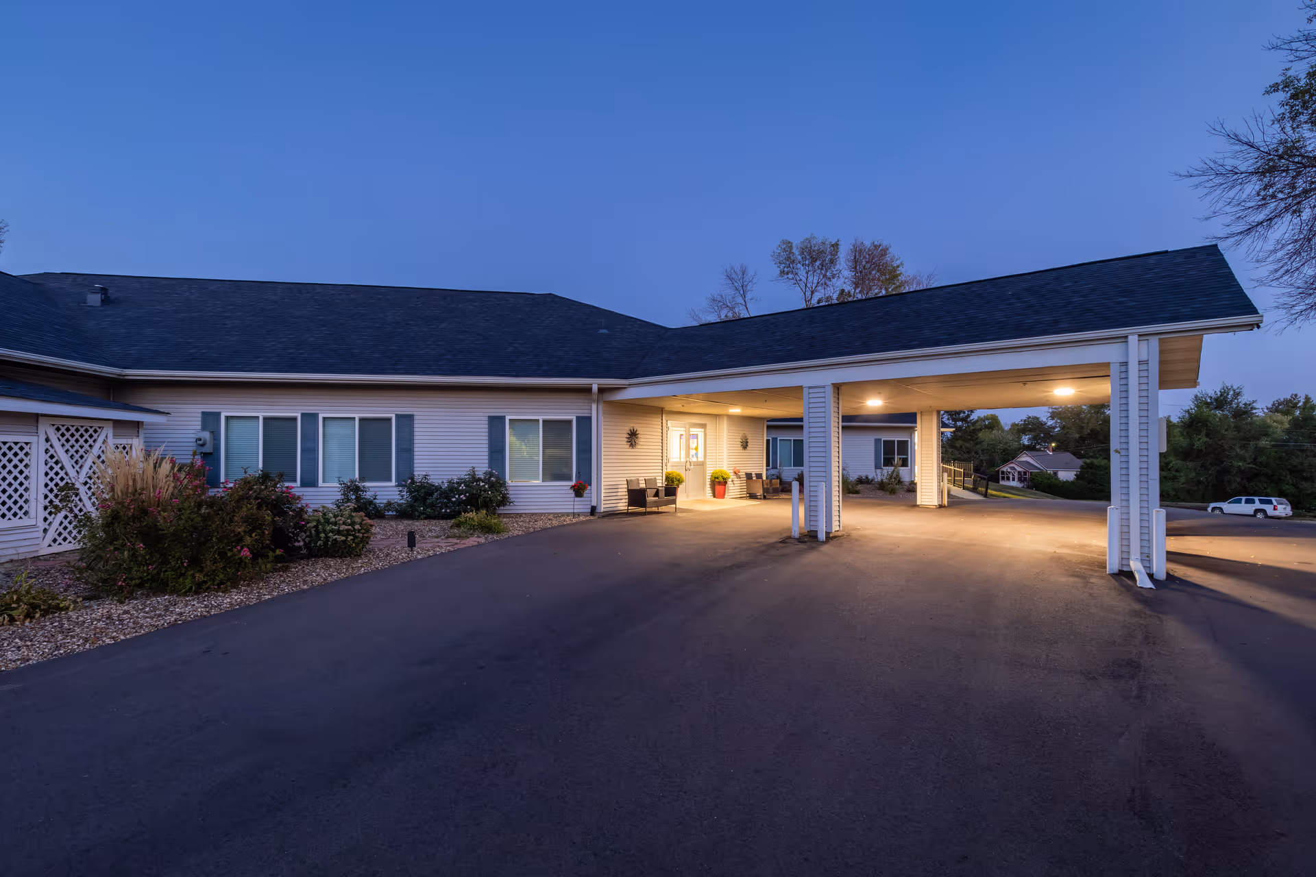 Exterior view of Corridor Crossing Place facility at dusk, showing a single-story building with a covered entrance driveway, illuminated by outdoor lights. The building has white siding, several windows with blue shutters, and landscaped bushes along the front. A white vehicle is parked in the background near trees and other houses.