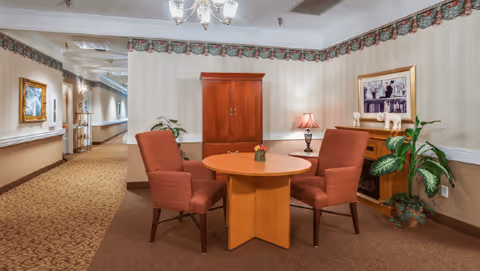 A hallway seating nook with a round wooden table surrounded by upholstered chairs, an armoire, potted plants, and framed artwork.