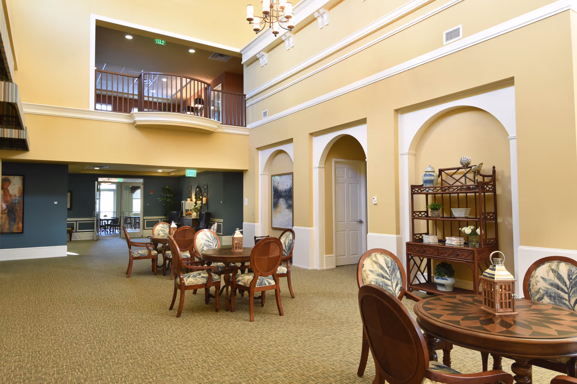 Spacious, well-lit communal dining area with round wooden tables and chairs, decorative shelving, and an upper balcony overlook.