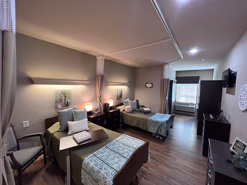 A shared senior living bedroom with two twin beds, nightstands and lamps, seating, and a window at the far end.