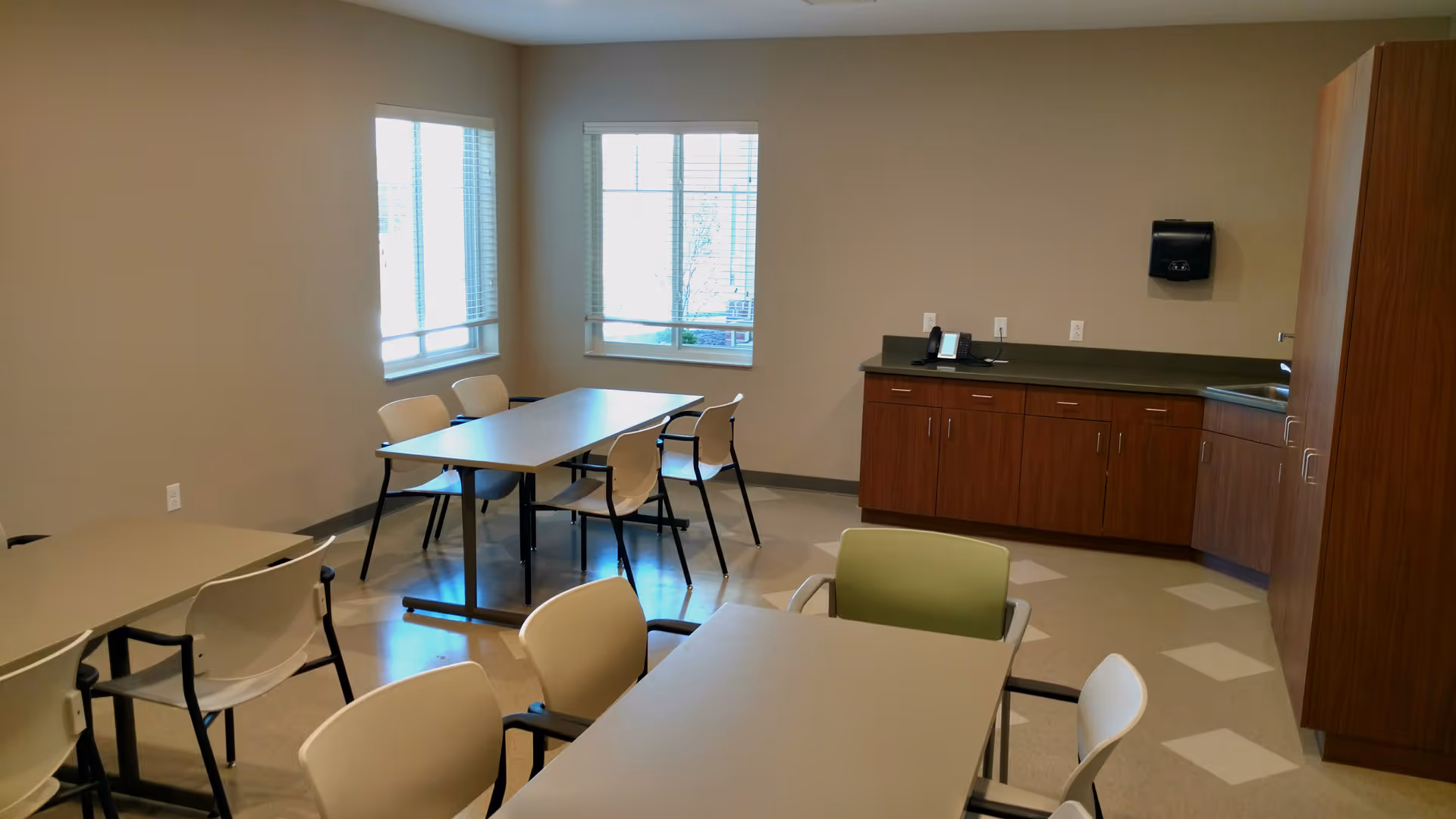A small dining area with several tables and chairs arranged around the room. There are two windows letting in natural light, and a wooden cabinet with a countertop and a paper towel dispenser on the wall. The room has a clean and simple design with beige walls and a tiled floor.