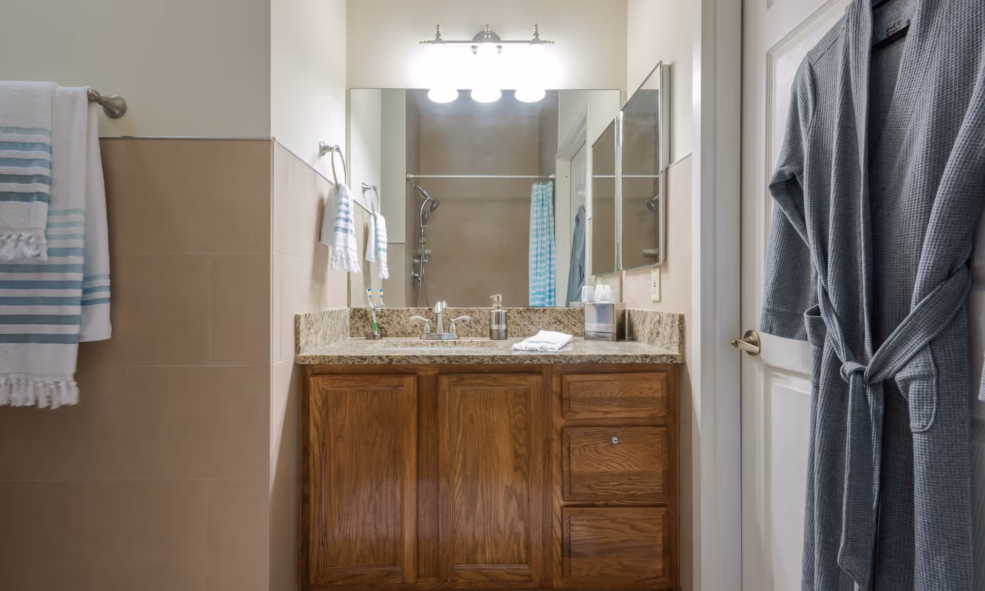 A bathroom vanity with a granite countertop, wooden cabinets, and a large mirror above. There are three light fixtures above the mirror. On the countertop, there is a soap dispenser, a tissue box, and a folded white towel. To the left, a towel rack holds two white and blue striped towels. On the right side, a gray bathrobe hangs on a door. In the reflection of the mirror, a shower with a blue and white striped curtain is visible.