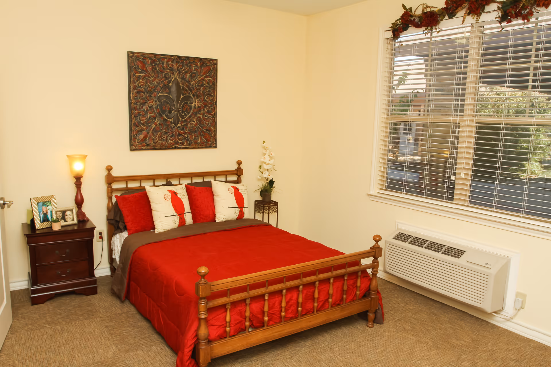 A bedroom with a wooden bed frame and a red bedspread. The bed has four pillows, two of which have a red bird design. There is a nightstand with a lamp and framed photos on the left side of the bed. A decorative wall hanging is above the bed. A window with blinds is on the right wall, and below it is a wall-mounted air conditioning unit. The room has beige walls and carpeted flooring.