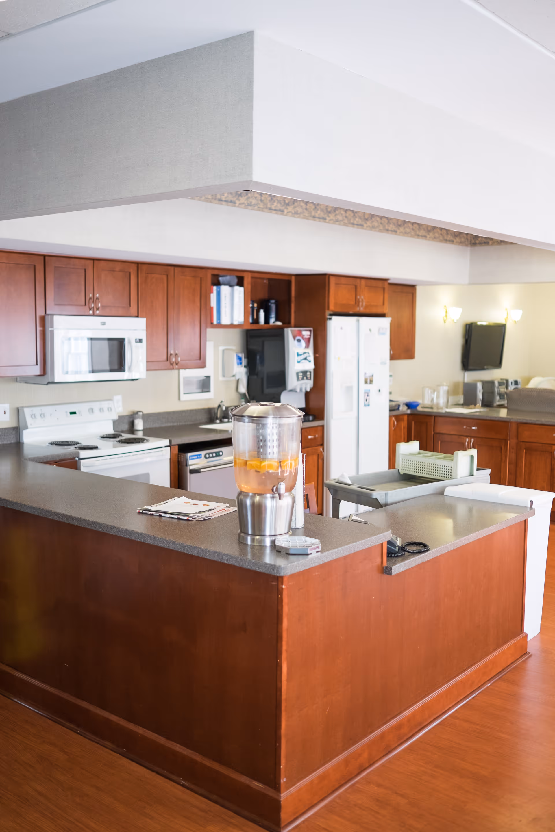 A clean and well-lit kitchen area with wooden cabinets, a white stove, microwave, refrigerator, and dishwasher. A countertop island with a beverage dispenser containing orange slices is in the foreground. The kitchen has a modern and organized appearance.