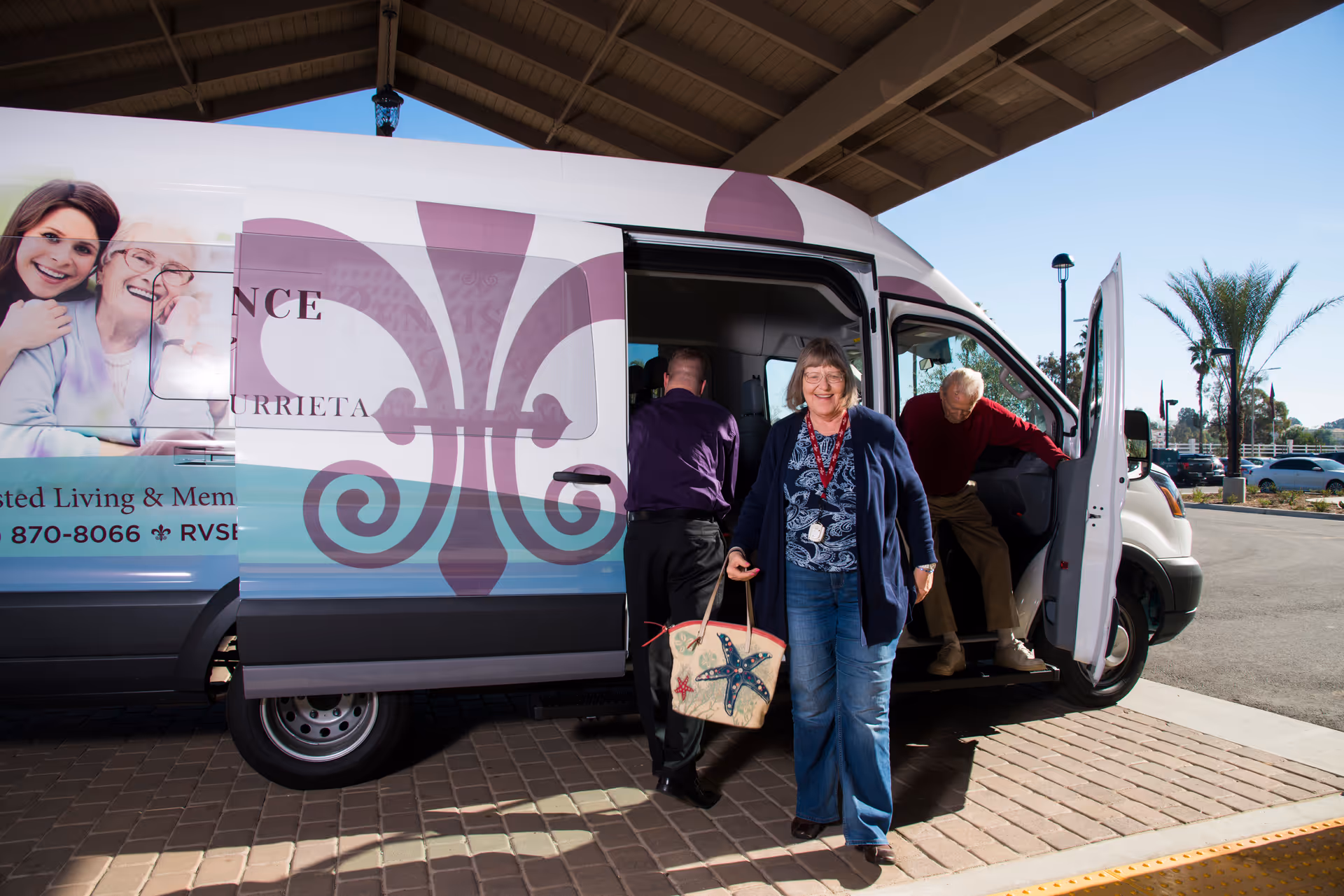 An elderly woman smiling and carrying a tote bag with starfish designs walks away from a white shuttle van with the Renaissance Village Murrieta logo. An elderly man is stepping out of the van, and a man in a purple shirt is standing inside the van. The scene is outdoors under a covered area with palm trees and parked cars in the background.