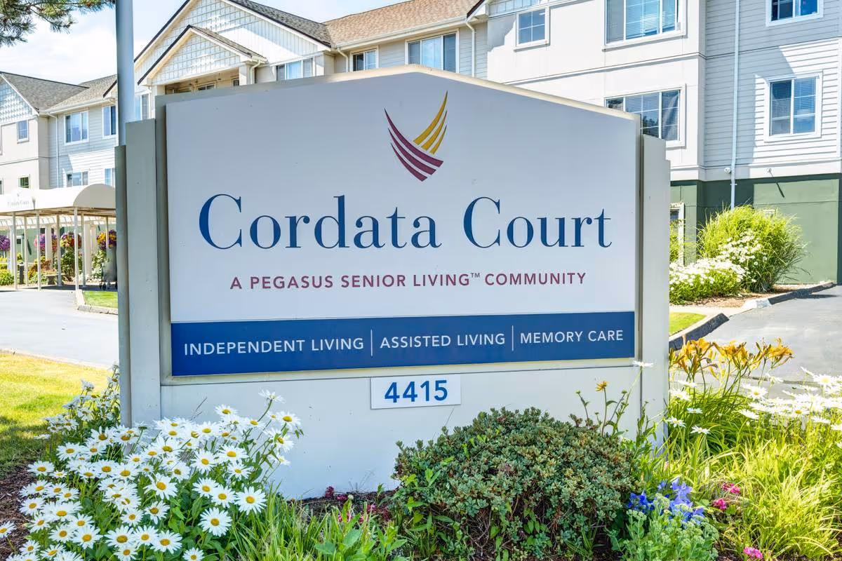 Outdoor view of a sign for Cordata Court, a Pegasus Senior Living community offering independent living, assisted living, and memory care. The sign is surrounded by flowers and greenery, with a multi-story building in the background.
