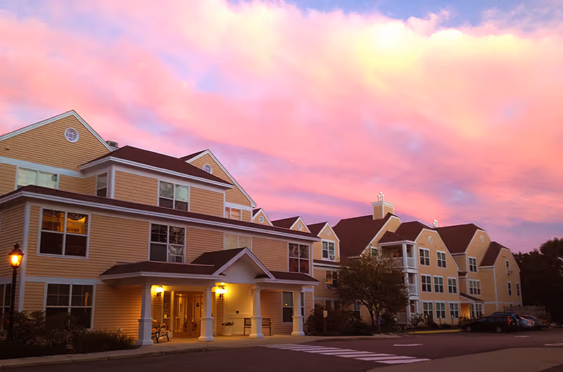 Exterior view of a large, multi-story senior living facility building at sunset with a pink and purple sky. The building has beige siding, multiple windows, and a covered entrance with lights on. There are some trees and parked cars visible near the building.