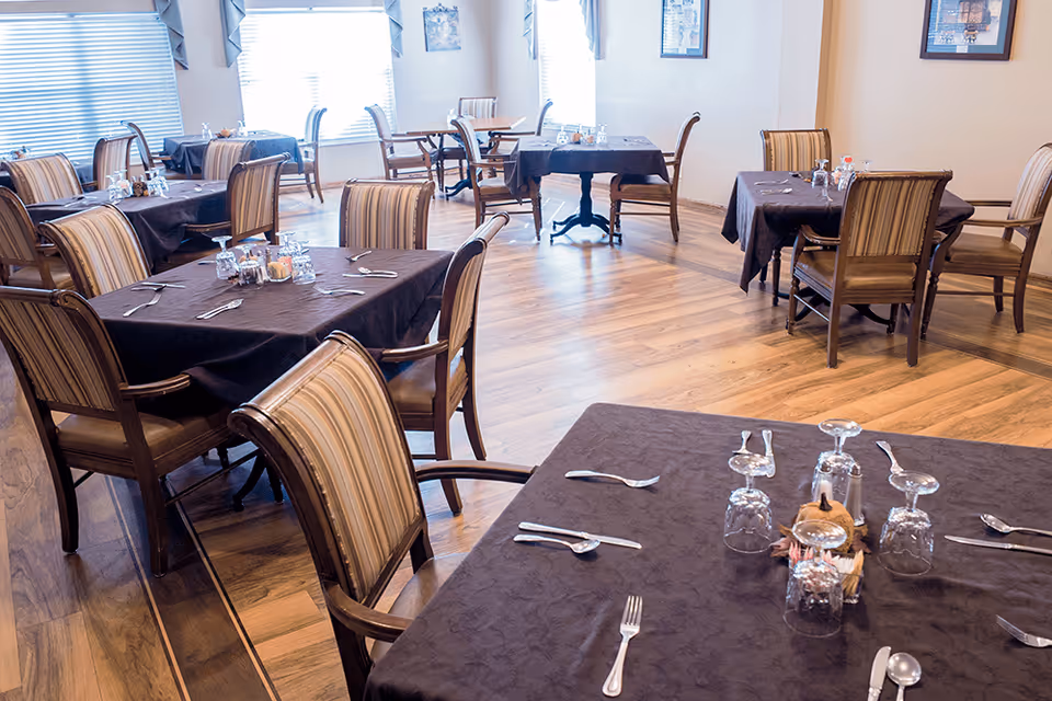 Dining room with multiple tables covered in dark tablecloths and set with utensils, glasses, and chairs on wood flooring.