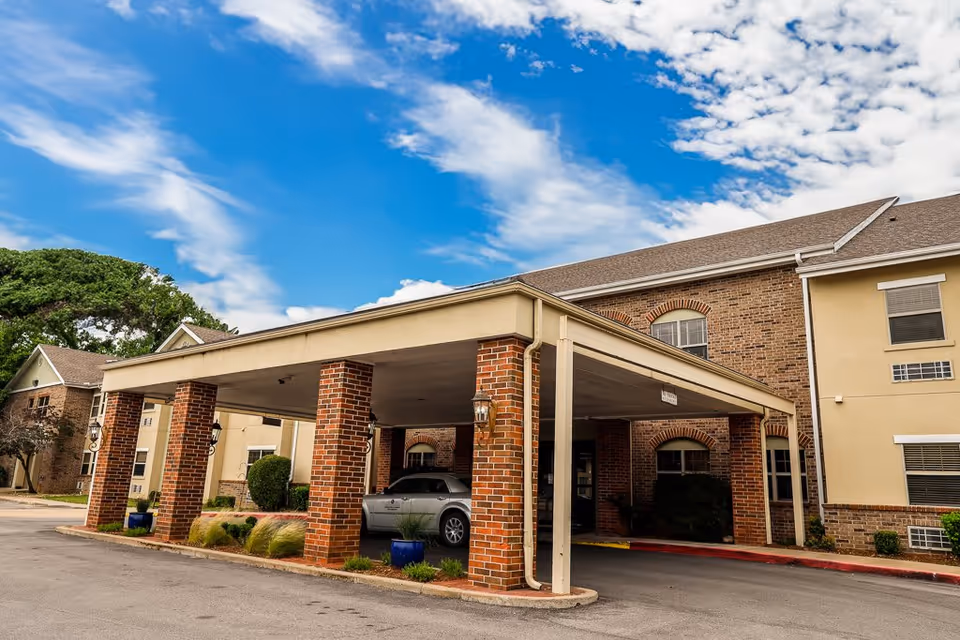 Exterior view of Aberdeen Heights Assisted Living facility showing a covered entrance with brick pillars, a parked car underneath, and a two-story building with brick and beige siding under a partly cloudy blue sky.