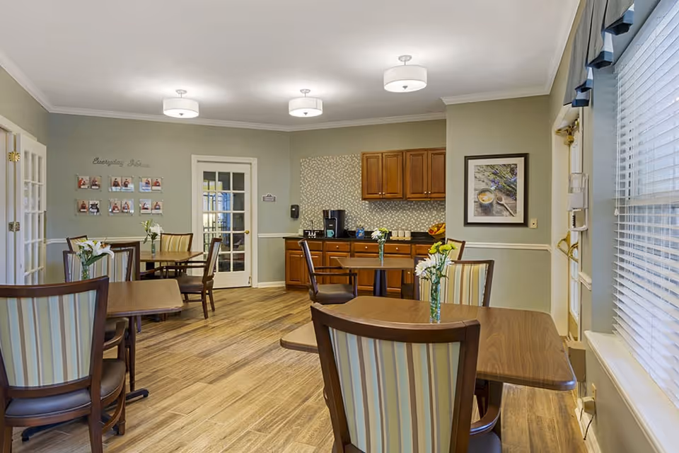 A dining room area in a senior living facility with wooden tables and striped cushioned chairs. Each table has a small vase with white flowers. The room has light green walls, wooden flooring, and a kitchenette with wooden cabinets and a coffee machine. There is a framed picture on the wall and a door with glass panels leading to another room.