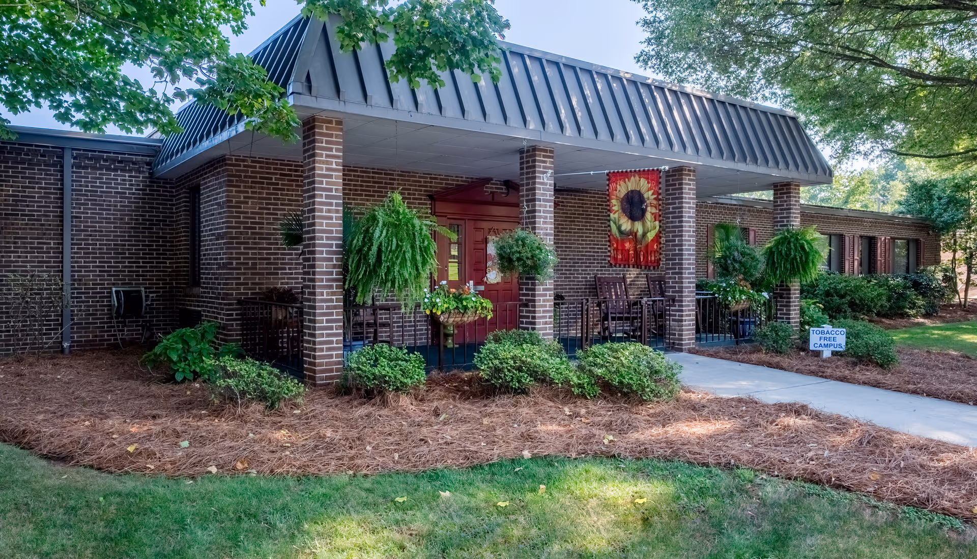Exterior view of a brick building with a covered entrance supported by brick columns. The entrance area features hanging plants, potted flowers, and outdoor chairs. A decorative banner with a sunflower hangs near the door. There is a small sign on the lawn that reads 'TOBACCO FREE CAMPUS'. The surrounding area has green grass, bushes, and trees providing shade.