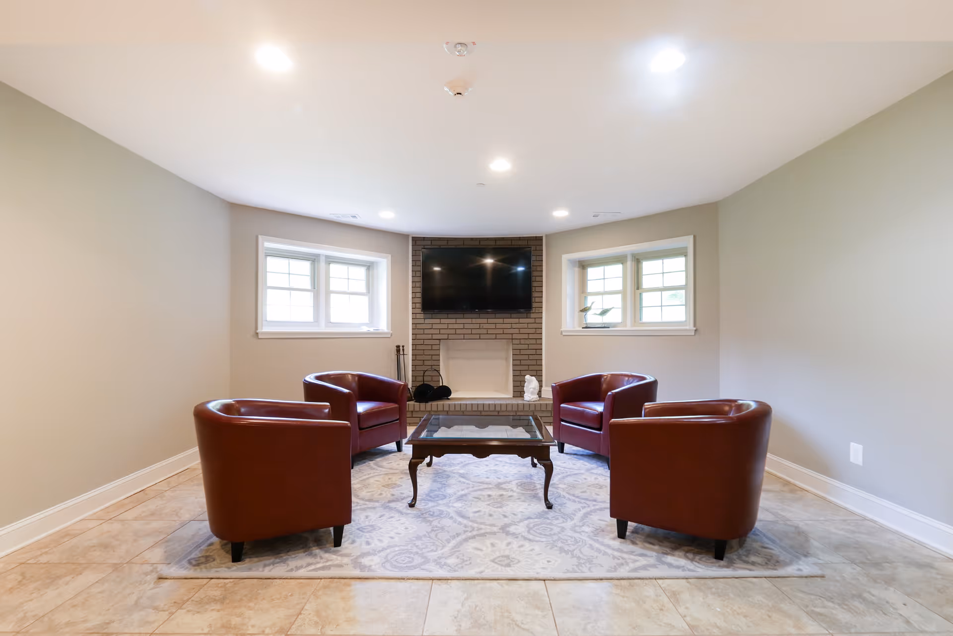 A cozy living room with four brown leather armchairs arranged around a glass coffee table on a patterned area rug. The room has beige tiled flooring, light gray walls, two windows on either side of a brick fireplace with a mounted flat-screen TV above it.