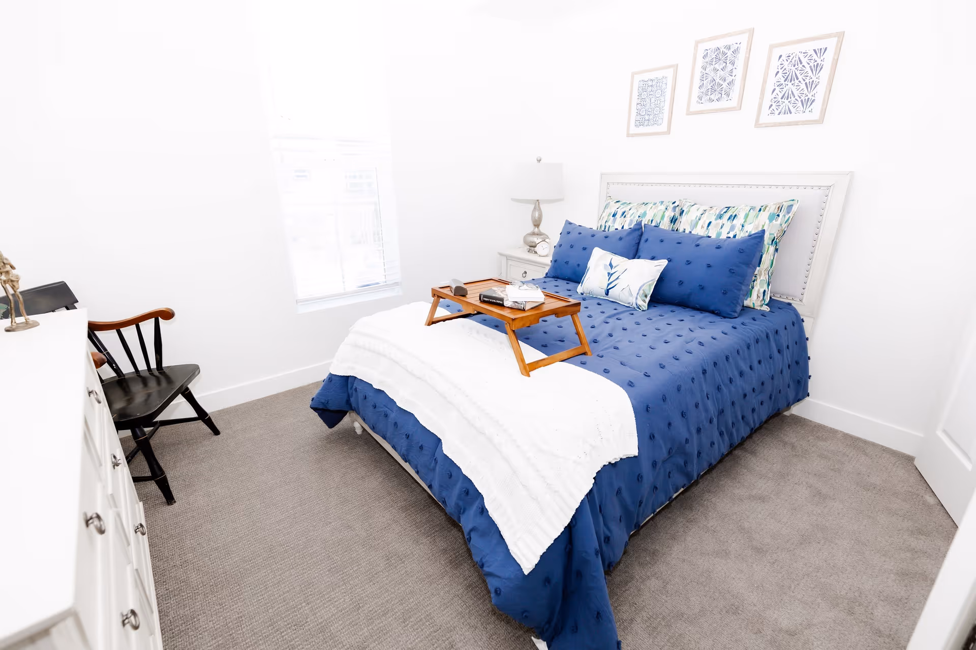 A bright bedroom with a bed dressed in a blue comforter and multiple pillows, including patterned and solid blue ones. A white throw blanket is draped across the foot of the bed, and a wooden tray with books and a cup is placed on the bed. There is a white nightstand with a lamp and clock beside the bed, a black wooden chair, a white dresser, and three framed abstract artworks on the wall above the bed. The room has a window with blinds letting in natural light and light gray carpeted flooring.