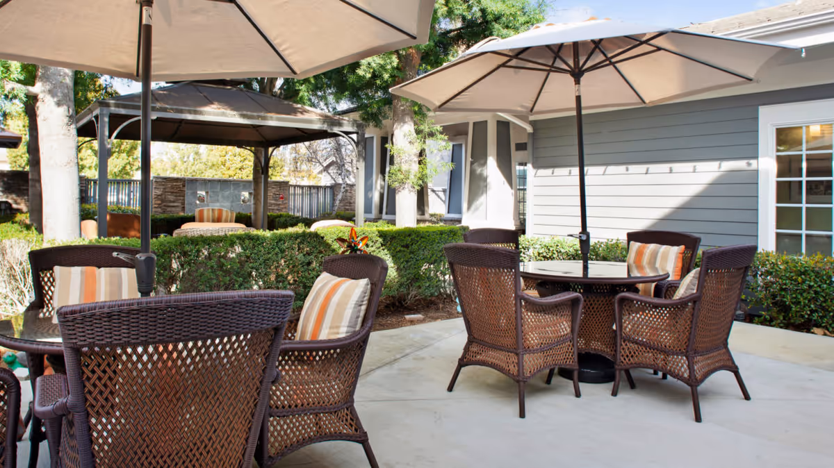 Outdoor patio area at a senior living facility with round tables and wicker chairs featuring striped cushions. Large umbrellas provide shade, and there is a gazebo in the background surrounded by greenery and trees. The building exterior is visible with gray siding and white-framed windows.