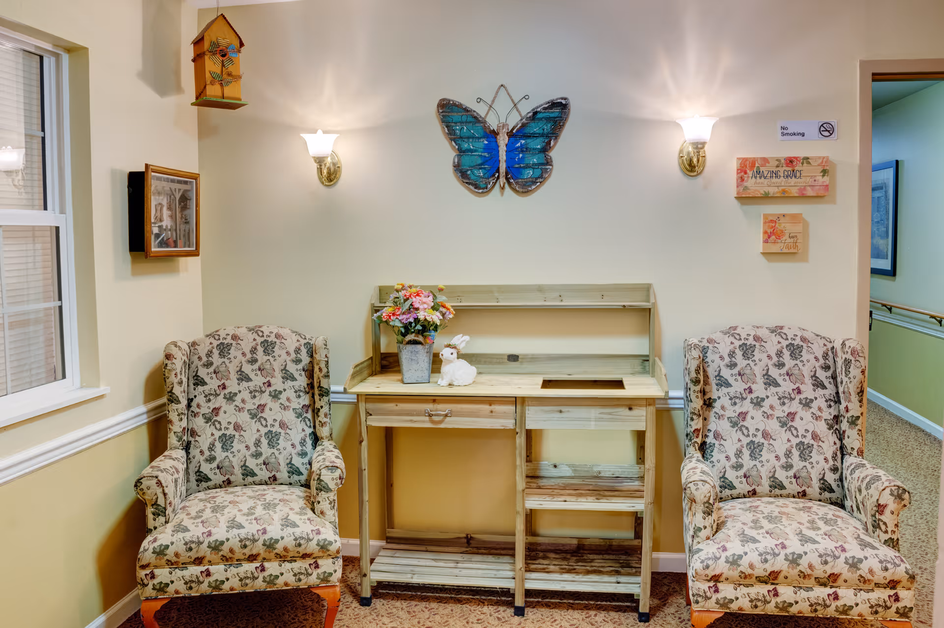 A cozy sitting area with two floral upholstered armchairs on either side of a wooden desk. The wall behind the desk features a large blue butterfly decoration, two wall sconces with lights, a birdhouse, and framed artwork. There are also small decorative signs on the wall, and a window is visible on the left side.