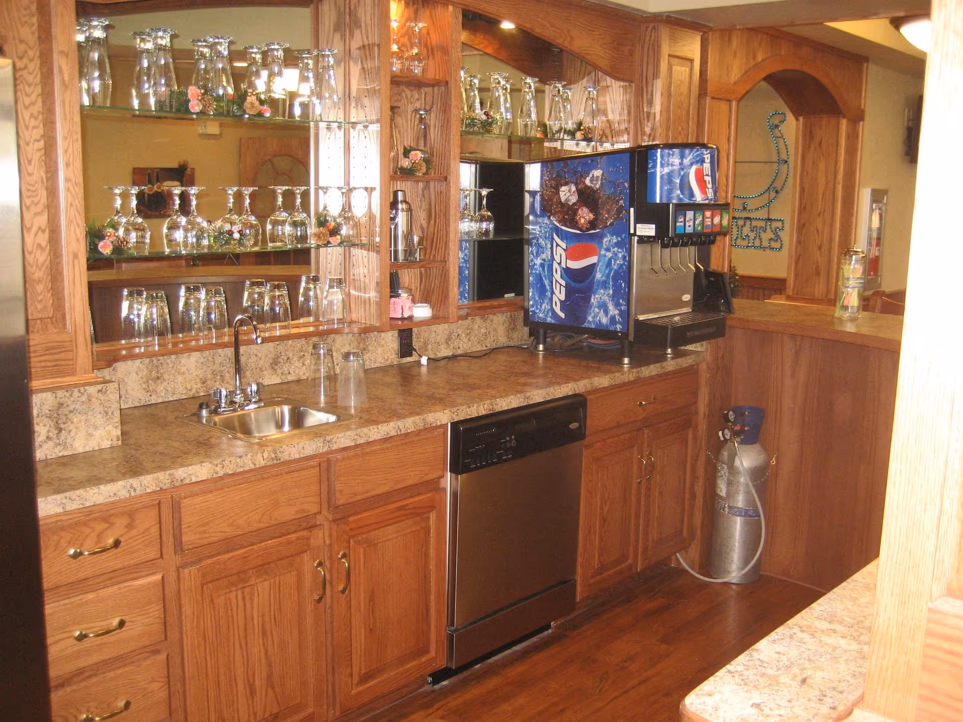 Interior view of a kitchen area with wooden cabinets, a countertop with a small sink, a dishwasher, and a Pepsi soda fountain machine. Glass shelves with various glassware are mounted on the wall above the counter. The floor is wooden, and there is a large metal tank on the floor near the soda machine.