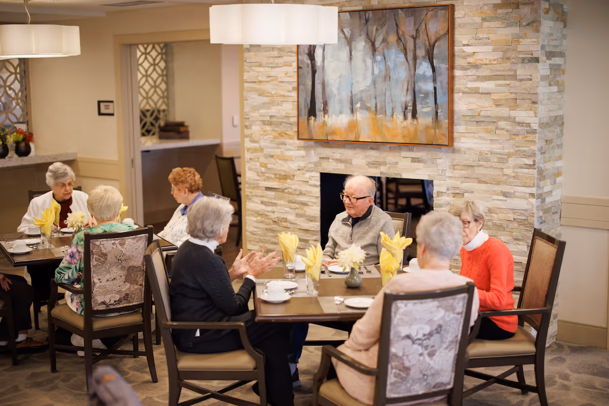 A group of elderly people sitting around tables in a dining area, engaged in conversation. The room features a stone accent wall with a painting of trees, modern light fixtures, and tables set with yellow folded napkins and white cups.