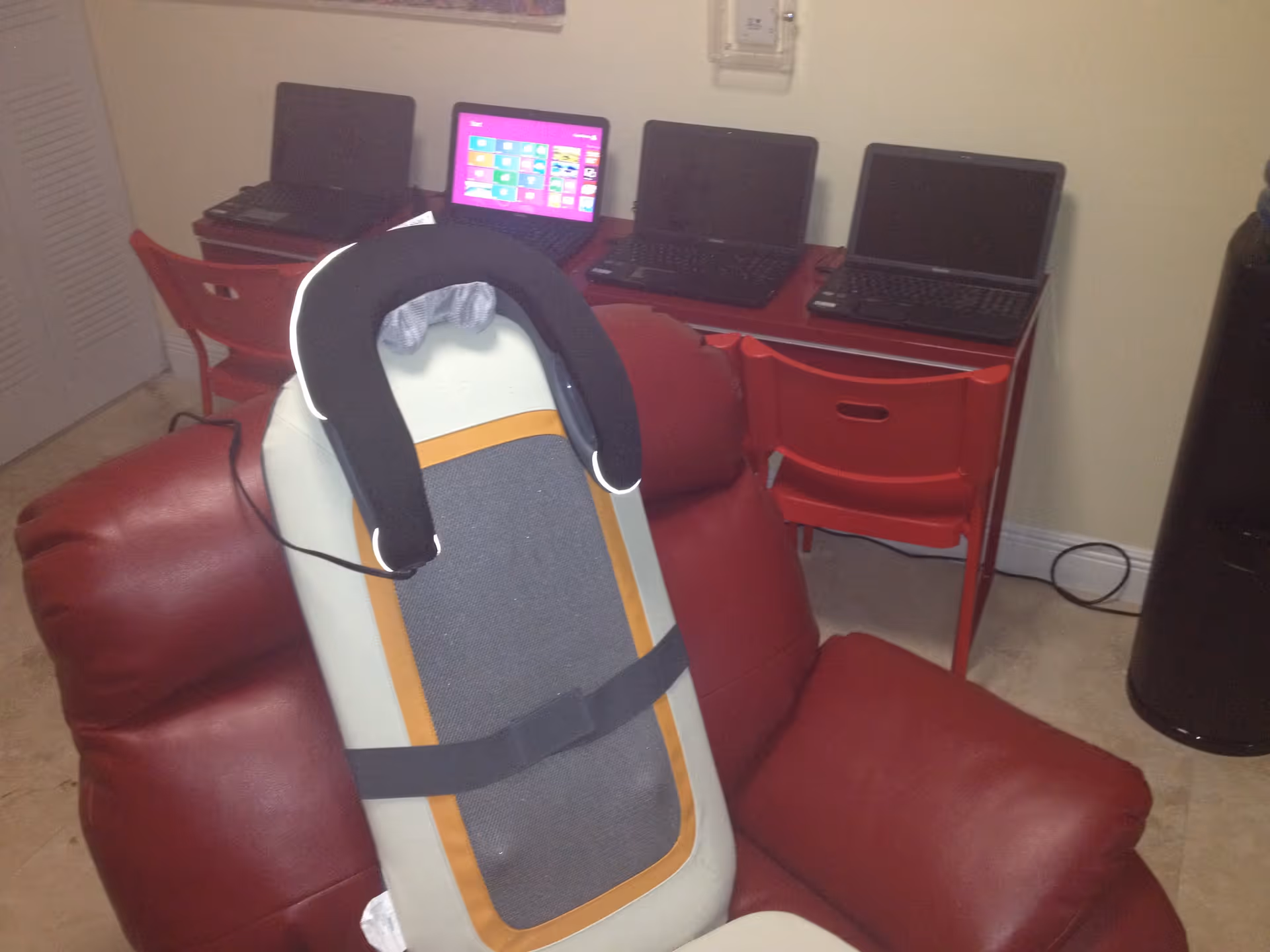 A red leather recliner chair with a massage cushion placed on it. Behind the chair, there is a red desk with three laptops on it and two red chairs. The room has beige walls and a tiled floor.
