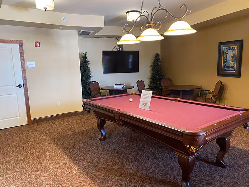 A recreational room with a red felt pool table in the center, a sign placed on the table, a wall-mounted flat screen TV, two small round tables with chairs, two artificial plants, a framed picture on the wall, and a ceiling light fixture with four lamps.