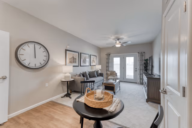 A bright living room in a senior living facility featuring a gray sofa with multiple cushions, a side table with a lamp, framed artwork on the wall, a round black table with a woven basket centerpiece, a TV on a wooden stand, and double glass doors leading outside. The room has a ceiling fan and light-colored walls and carpet.