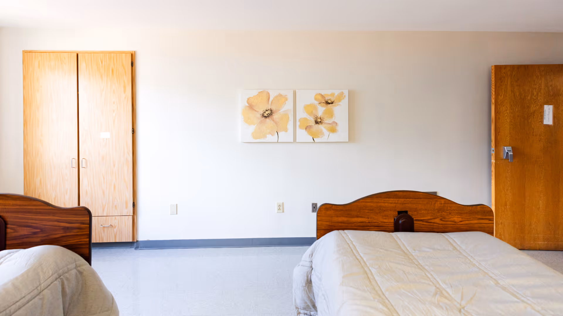 Simple shared bedroom with two twin beds with wooden headboards, a wardrobe, a closed door, and floral wall art on a pale wall.