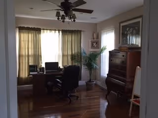 A cozy interior room with wooden flooring featuring a desk with a computer and office chair near a window with beige curtains. A ceiling fan with lights is mounted above. There is a tall potted plant next to a window with sheer white curtains, and a wooden piano with a framed picture above it is placed against the wall.