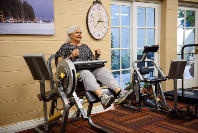 An elderly woman with gray hair is exercising on a seated leg press machine in a fitness room with large windows and a wall clock. The room has beige walls and a striped carpet floor.