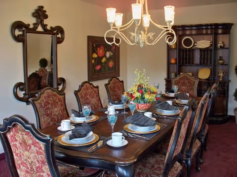 A formal dining room with a long wooden table set for eight people. The table is arranged with plates, cups, glasses, and black napkins. There are ornate chairs with red and gold upholstery around the table. A chandelier with six lights hangs above the table. A large decorative mirror and a floral painting are on the wall, and a dark wooden cabinet with shelves and folded linens is in the corner.