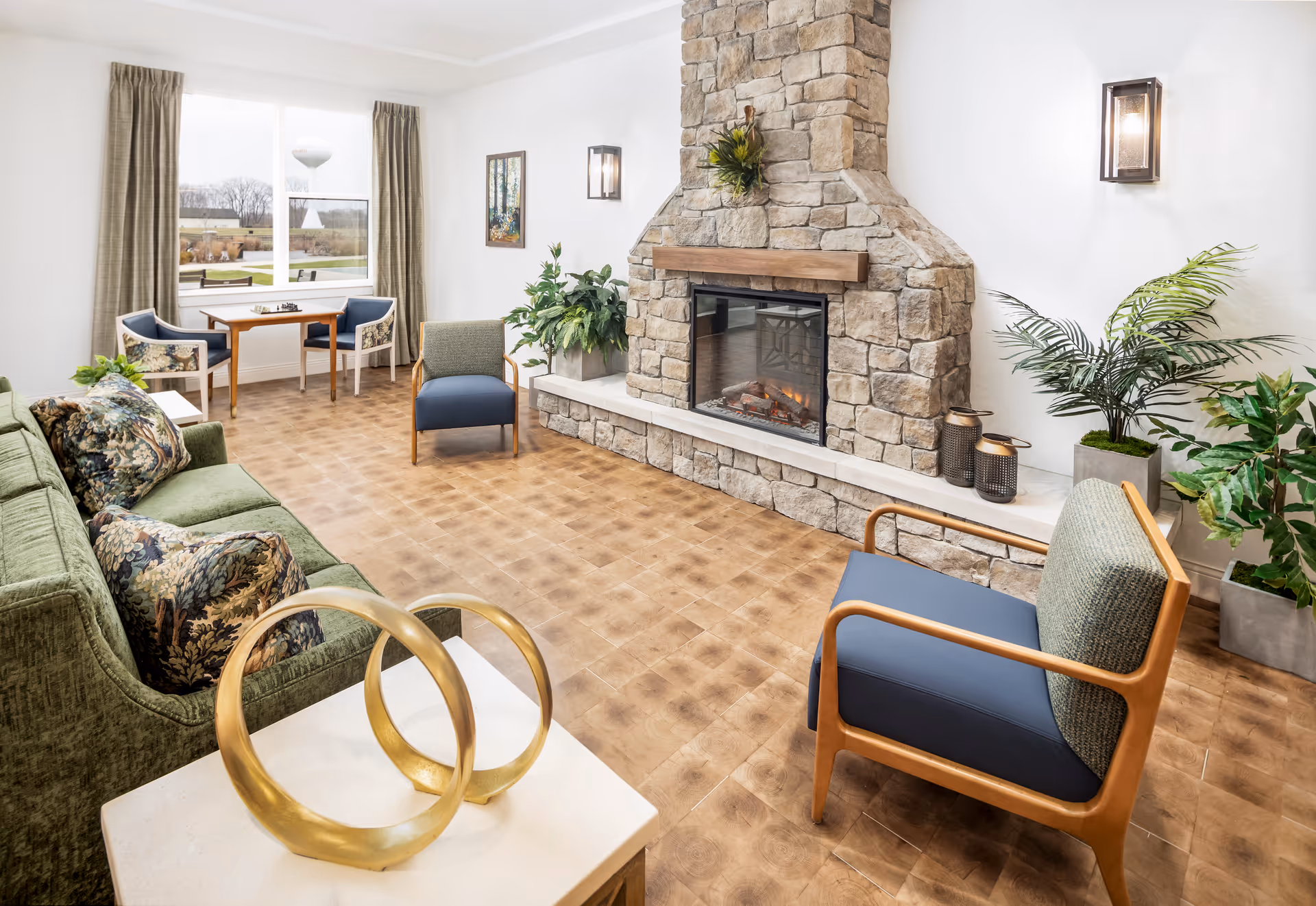 Bright common living room with a stone fireplace, sofa, chairs, plants, and a small table by the window.