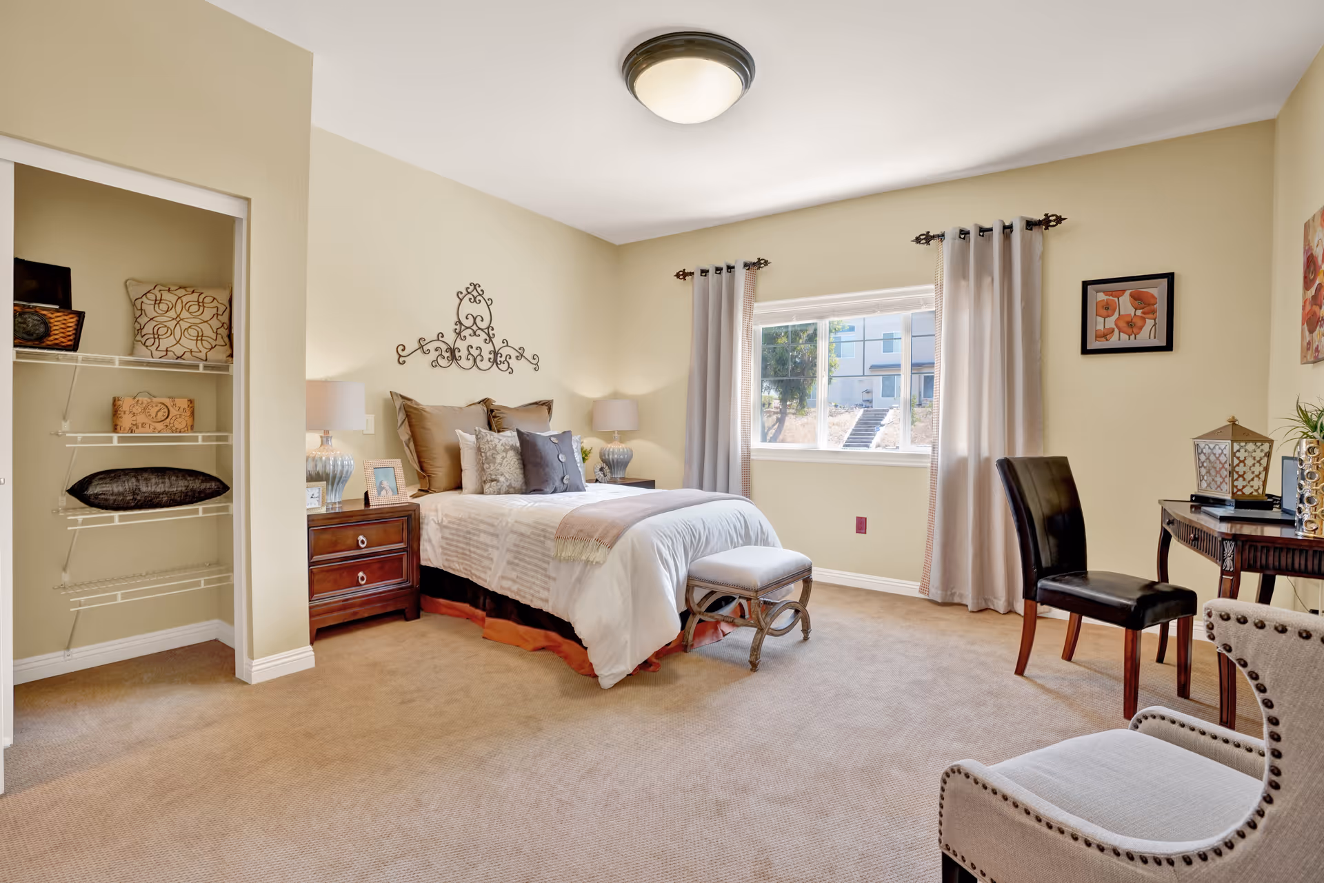 A bright and spacious bedroom with a neatly made bed featuring multiple pillows and a decorative metal wall art above the headboard. There are two bedside tables with lamps, a small bench at the foot of the bed, a window with curtains letting in natural light, a closet with shelves holding pillows and baskets, a wooden desk with a chair, and an upholstered armchair in the foreground.