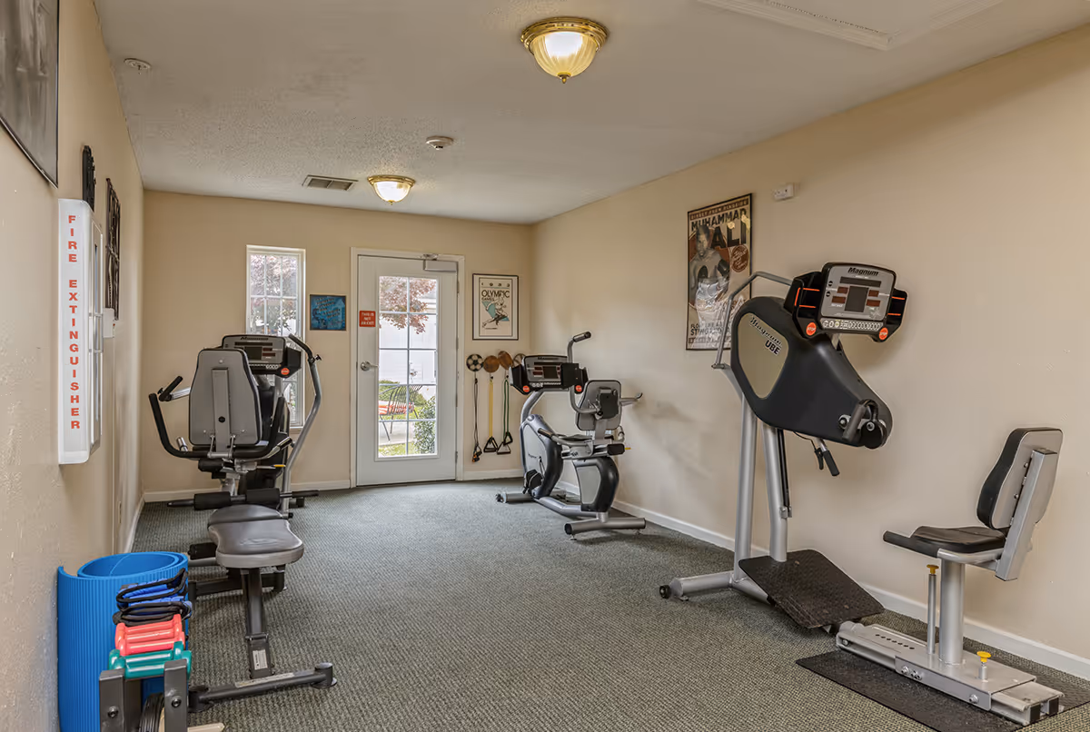 A small fitness room with exercise equipment including two recumbent bikes, a strength training machine, and various small weights and exercise mats. The room has beige walls, carpeted floor, ceiling lights, and a door with a window leading outside. Posters and exercise bands are mounted on the walls.