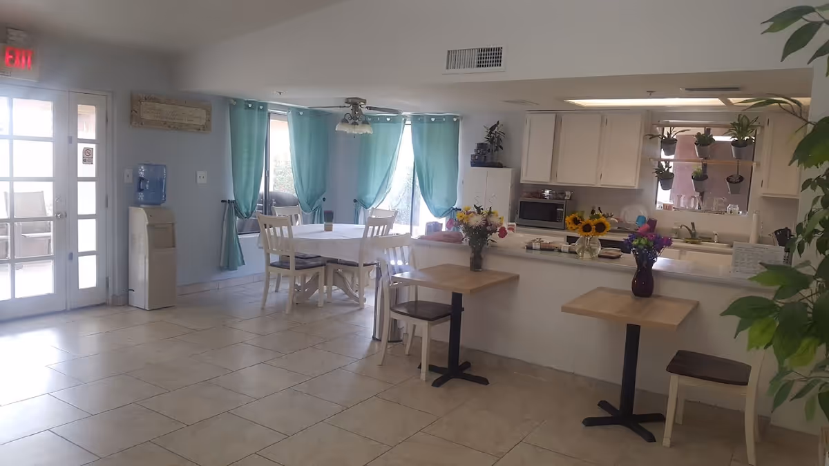 A bright and clean dining area in a care home with a round table surrounded by white chairs near windows with teal curtains. There are two small square tables with single chairs and flower vases on them. The kitchen area is visible in the background with white cabinets, a microwave, a sink, and potted plants on a shelf above the sink. A water cooler is near the exit door on the left side.