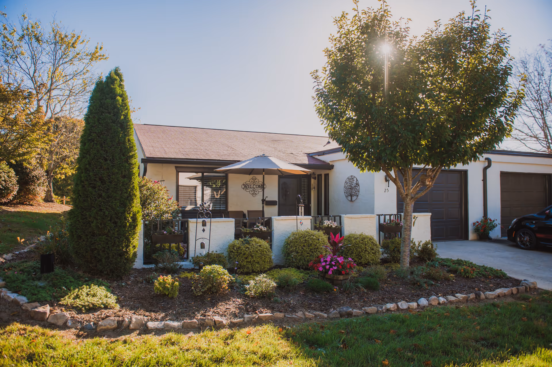 Front exterior of a single-story retirement community unit with a landscaped garden, small patio umbrella, and attached garage.