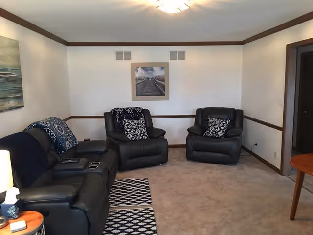 A cozy living room with three black leather recliners, each with patterned cushions. The room has beige carpet, white walls with wooden trim, and a ceiling light fixture. There are two framed pictures on the walls and a small wooden side table with a lamp and tissues.