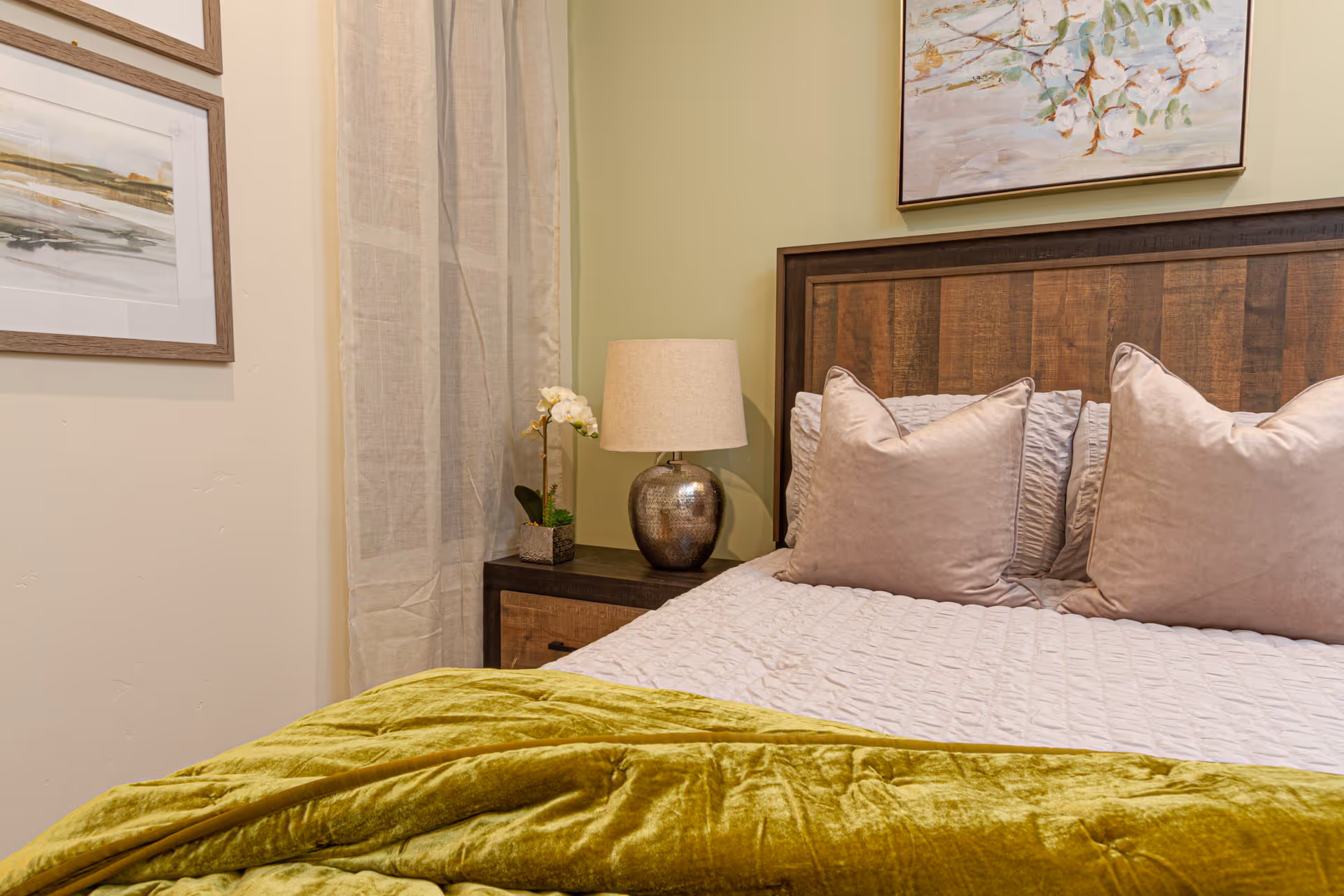 A cozy bedroom corner featuring a wooden headboard with two large beige pillows and a white quilted bedspread. A green velvet blanket is draped at the foot of the bed. Next to the bed is a dark wooden nightstand with a silver textured lamp and a small potted orchid. The walls are decorated with framed artwork and light beige curtains.