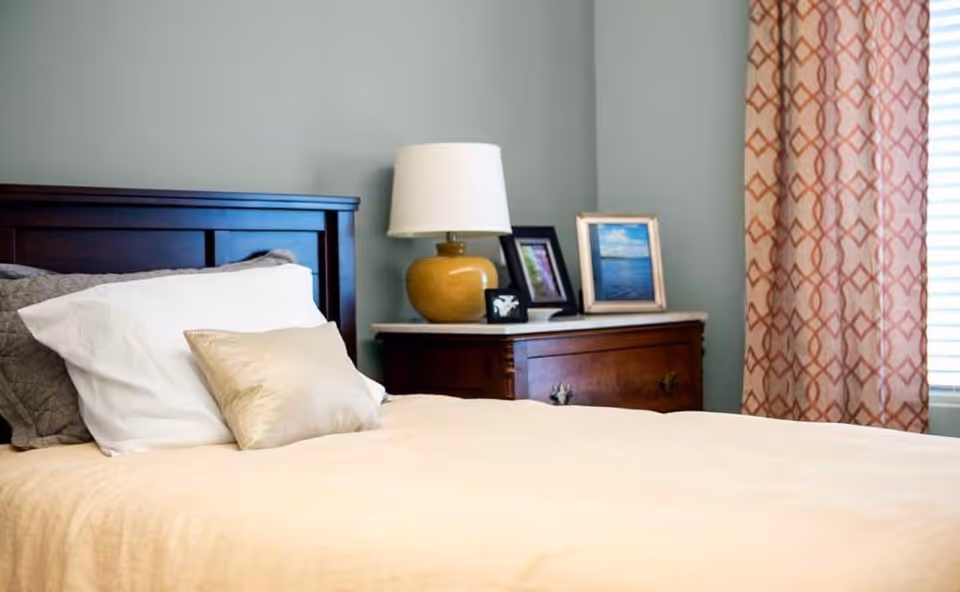 A neatly made bed with white and beige pillows in a bedroom. Next to the bed is a wooden nightstand with a yellow lamp and three framed pictures. A window with patterned curtains is visible on the right side.