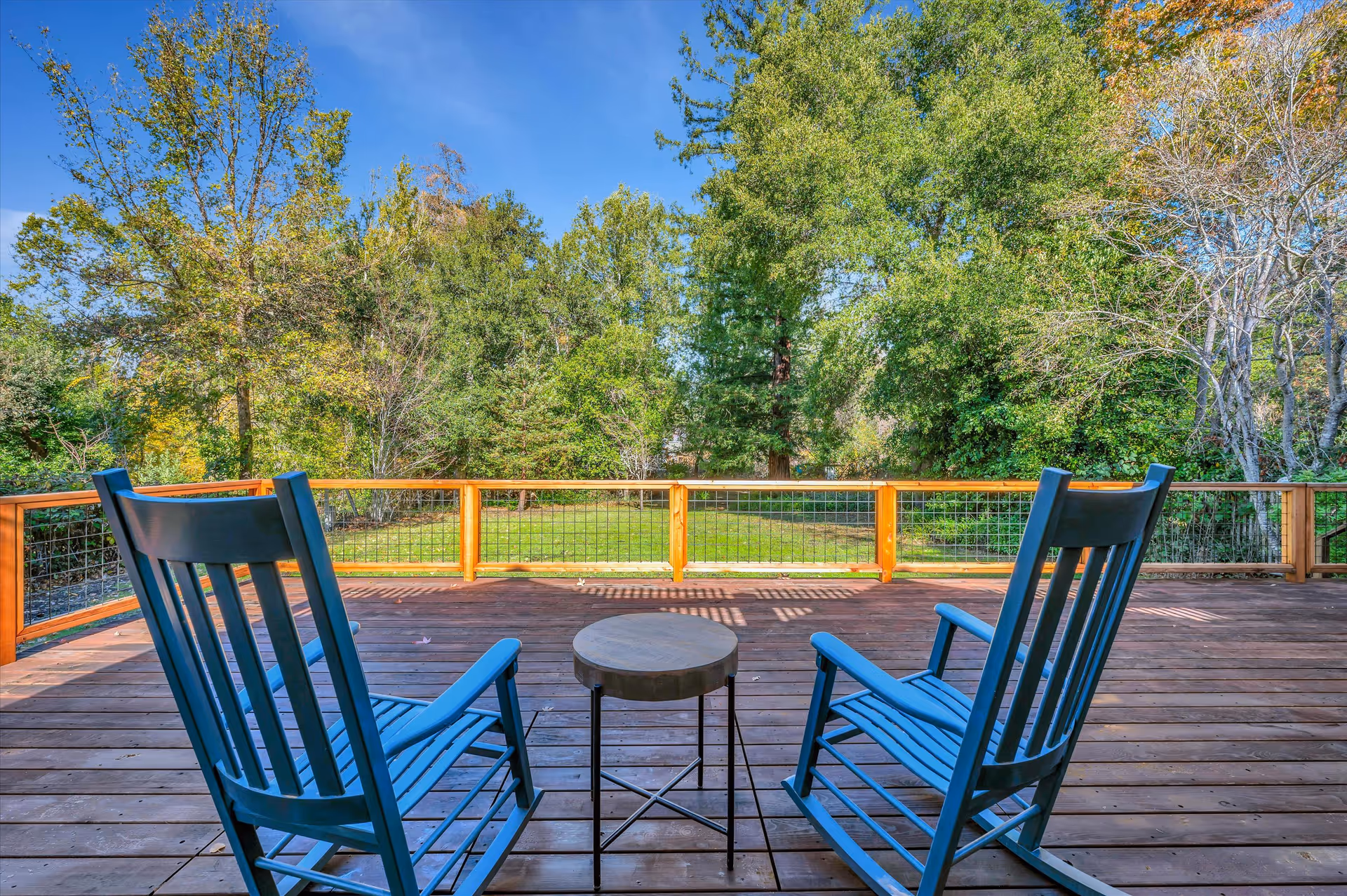 Two blue rocking chairs and a small round table on a wooden deck overlooking a green lawn and trees under a clear blue sky.