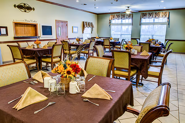 A bright dining room with multiple tables covered in brown tablecloths, each set with beige napkins, silverware, glassware, and floral centerpieces. The room has large windows with valances, tiled floors, and light green walls with decorative trim.