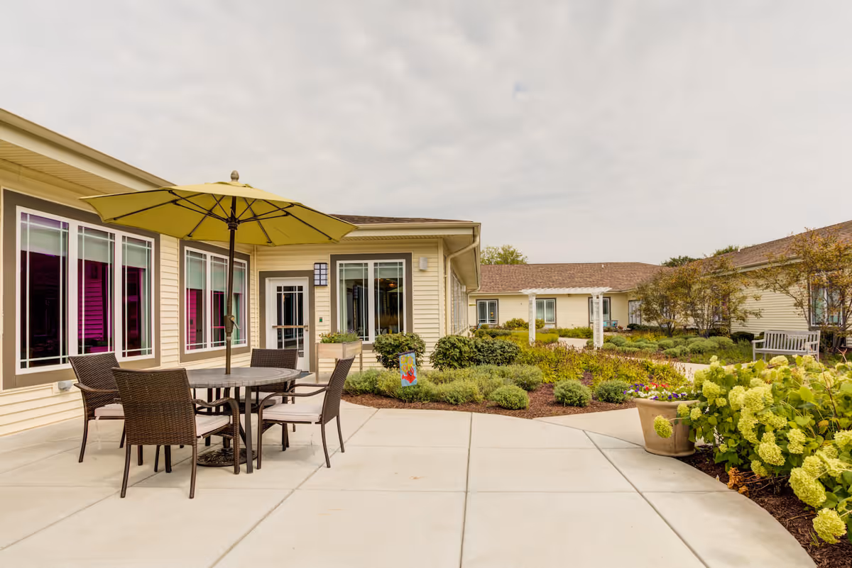 Outdoor patio area at Arbor Terrace Naperville featuring a round table with four wicker chairs and a large green umbrella. The patio is surrounded by well-maintained garden beds with various shrubs and flowers, and the building exterior is light yellow with multiple windows.