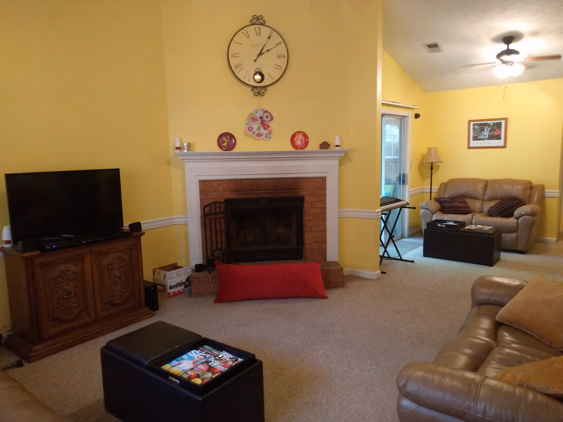 A cozy living room with yellow walls featuring a brick fireplace with a white mantel, a large clock above it, and decorative plates. There are two leather sofas with cushions, a wooden TV stand with a flat-screen TV, a black ottoman with magazines, a floor lamp, and a framed picture on the wall. A sliding glass door leads to an outdoor area.